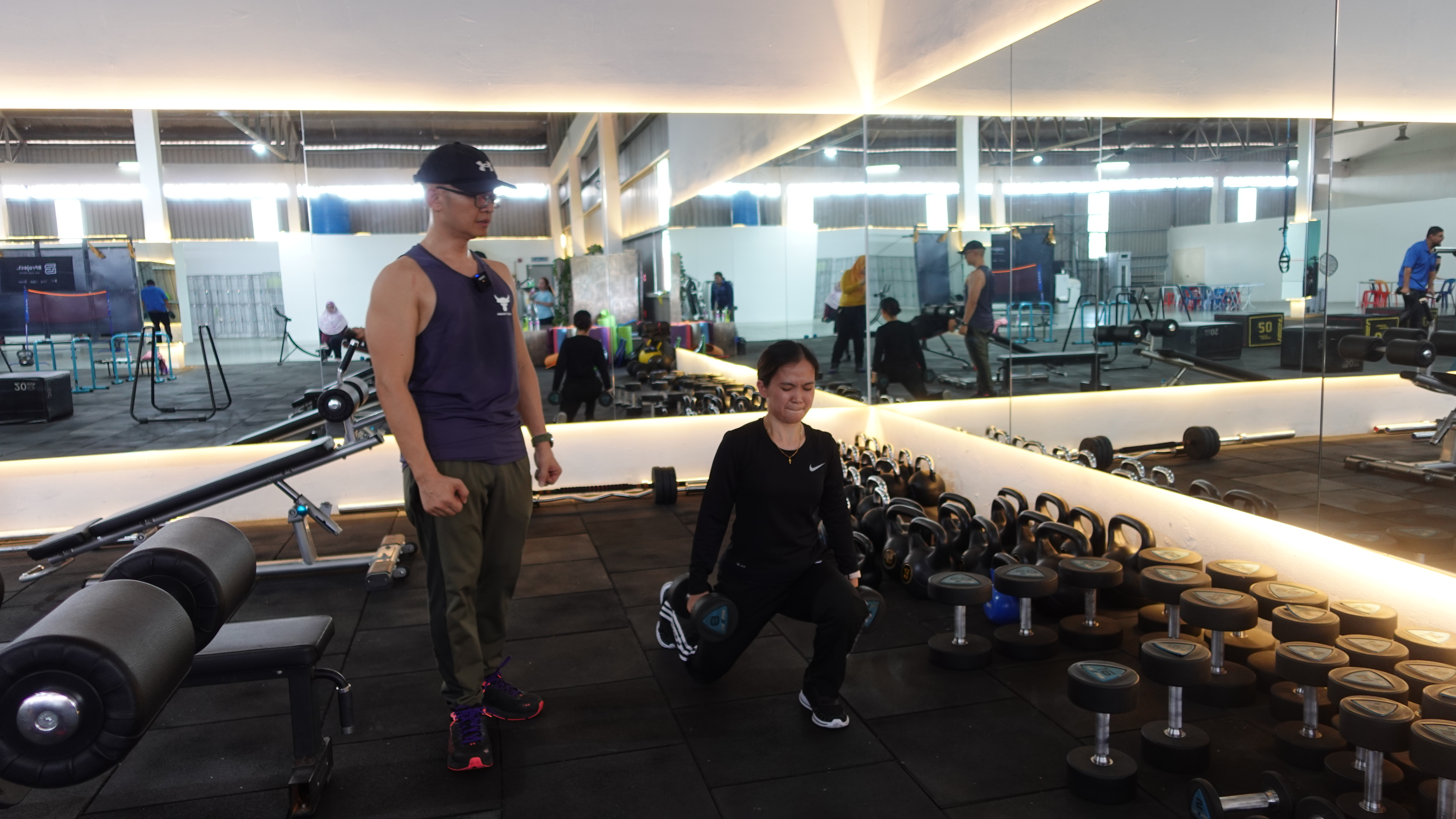 Coach Ho observing a participant performing a dumbbell lunge in a gym